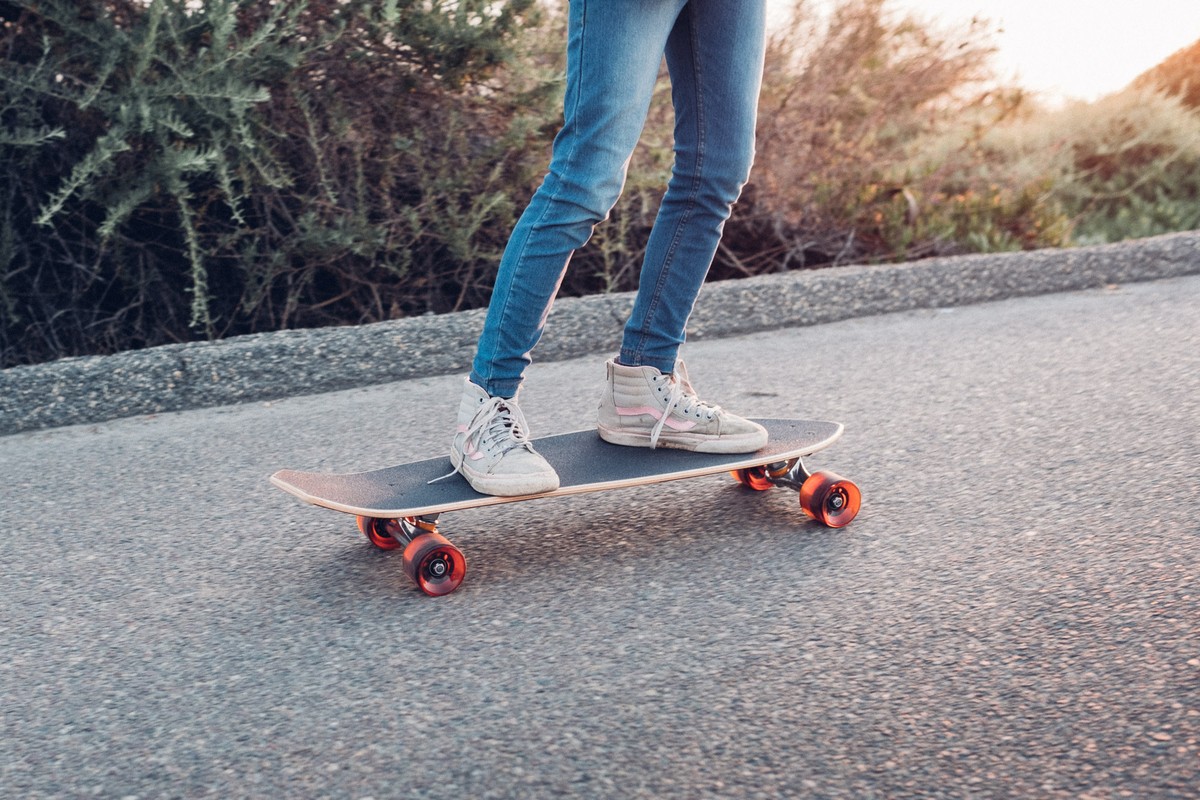 Close-up of legs in blue jeans and light high-top sneakers riding a skateboard on an asphalt road with foliage background.