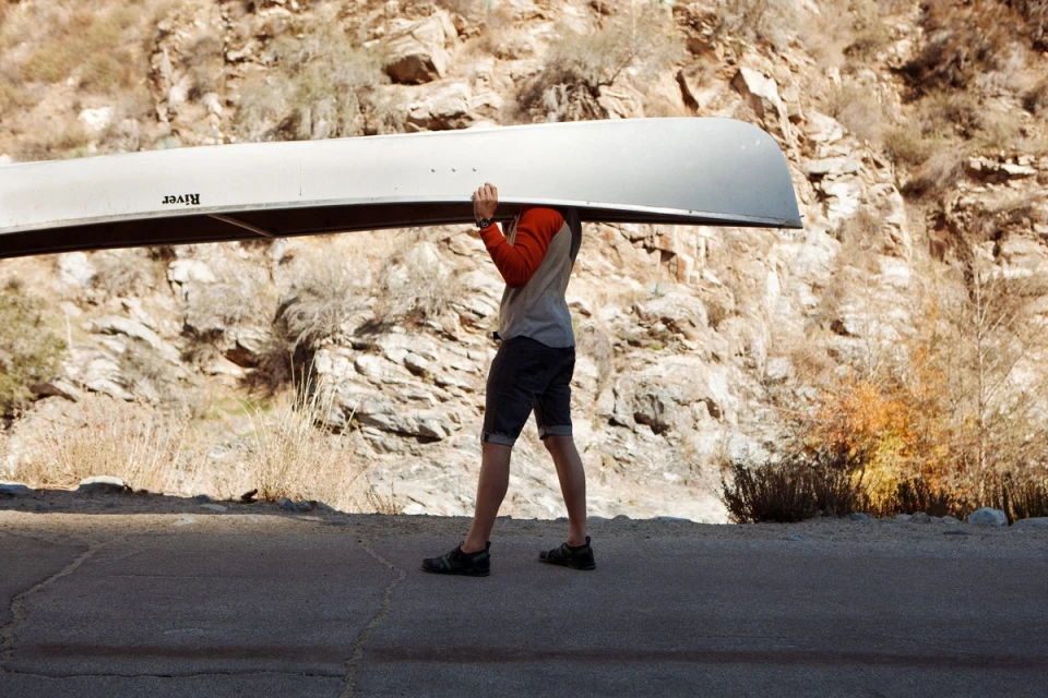Person carrying a canoe overhead on a road next to a rocky hillside. Outdoor shot, neutral tones, sunny day, riverbank setting.