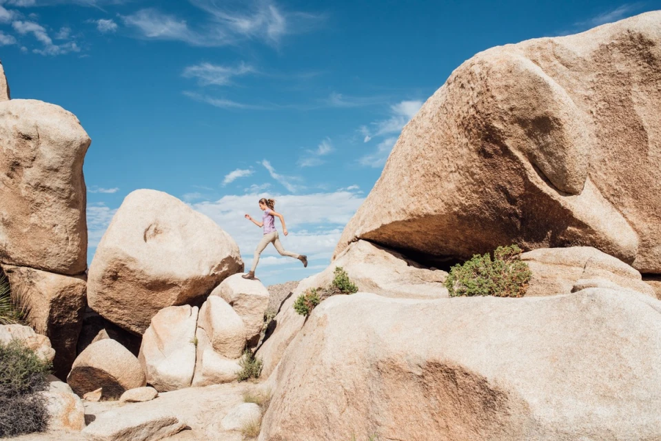 Woman jumping between large boulders in a desert landscape under a clear blue sky, capturing a moment of outdoor adventure.