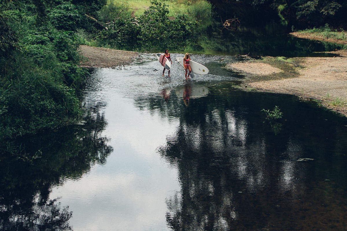 Two women in swimwear wade through a shallow river carrying surfboards, surrounded by lush green trees and foliage on a sunny day.