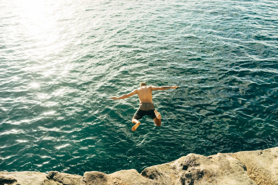Adult male jumps into turquoise ocean water from a rocky cliff on a sunny day with arms outstretched for watersport adventure.