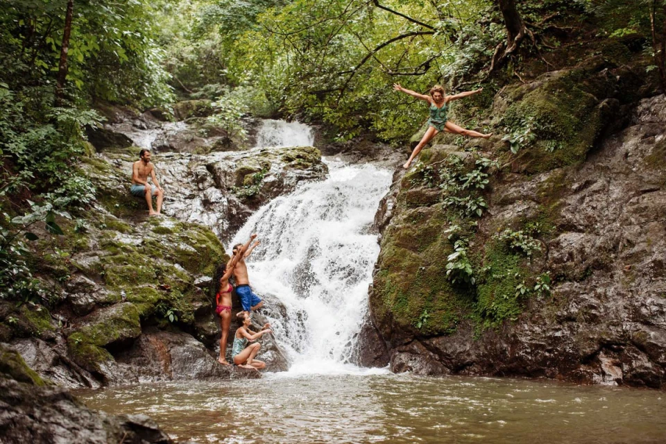 A group of people enjoys a refreshing day at a cascading waterfall surrounded by lush green trees and rocks in a tropical landscape.