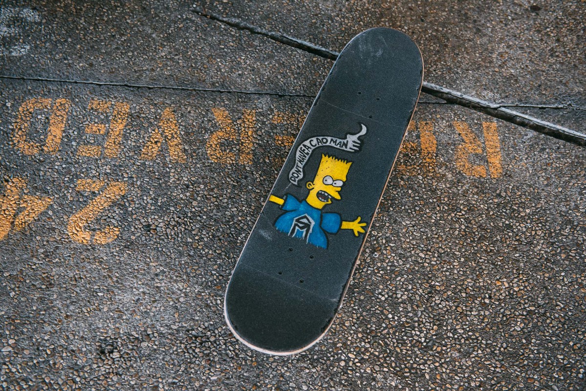 Overhead shot of black skateboard with Bart Simpson graphic lying on concrete with yellow painted text, showcasing street skateboarding culture.