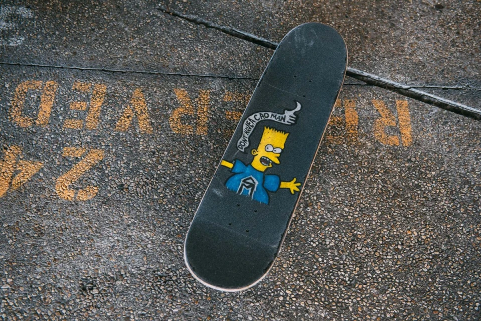 Overhead shot of black skateboard with Bart Simpson graphic lying on concrete with yellow painted text, showcasing street skateboarding culture.