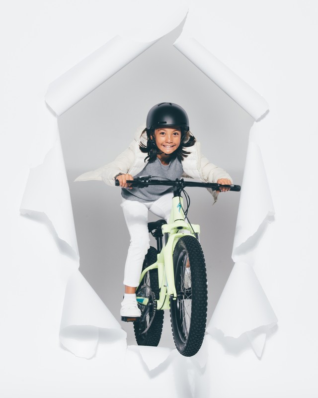 A young child wearing a helmet is riding a bike towards the viewer, framed by a white torn paper hole on a light grey background.