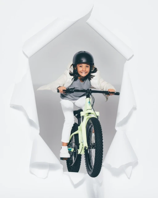 A young child wearing a helmet is riding a bike towards the viewer, framed by a white torn paper hole on a light grey background.