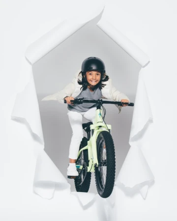 A young child wearing a helmet is riding a bike towards the viewer, framed by a white torn paper hole on a light grey background.