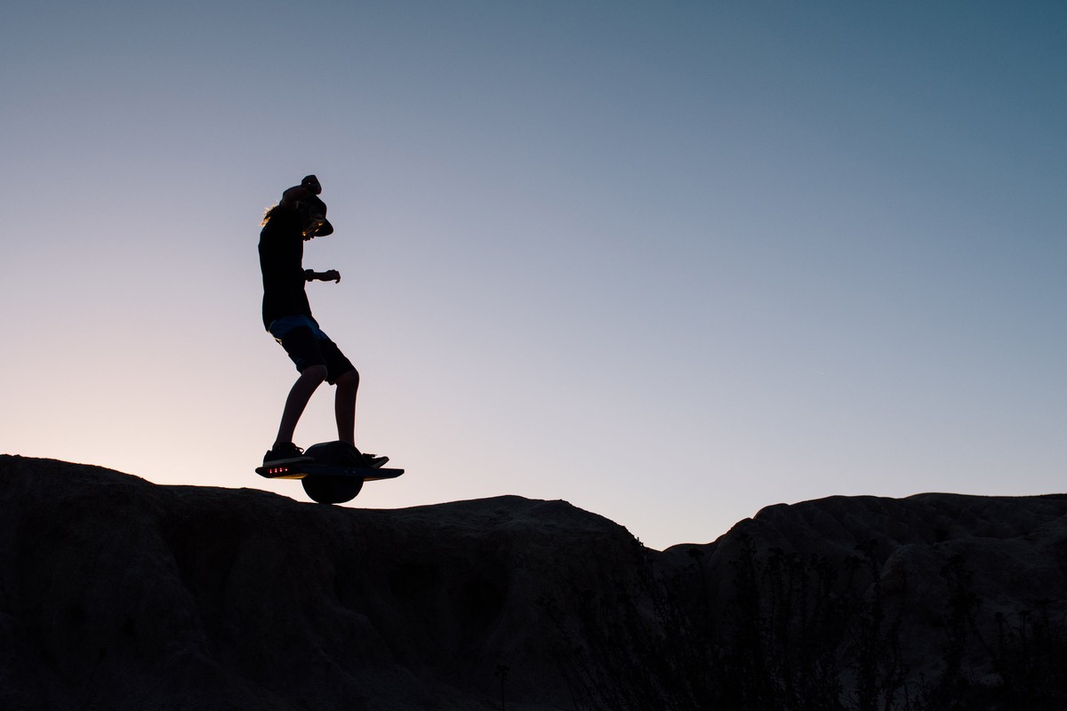 Silhouette of a man riding a Onewheel electric skateboard on a hill at sunset, against a gradient blue sky, outdoor environment.