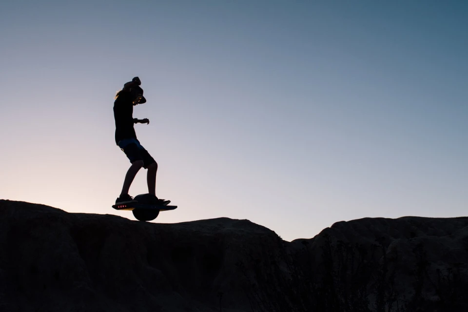 Silhouette of a man riding a Onewheel electric skateboard on a hill at sunset, against a gradient blue sky, outdoor environment.