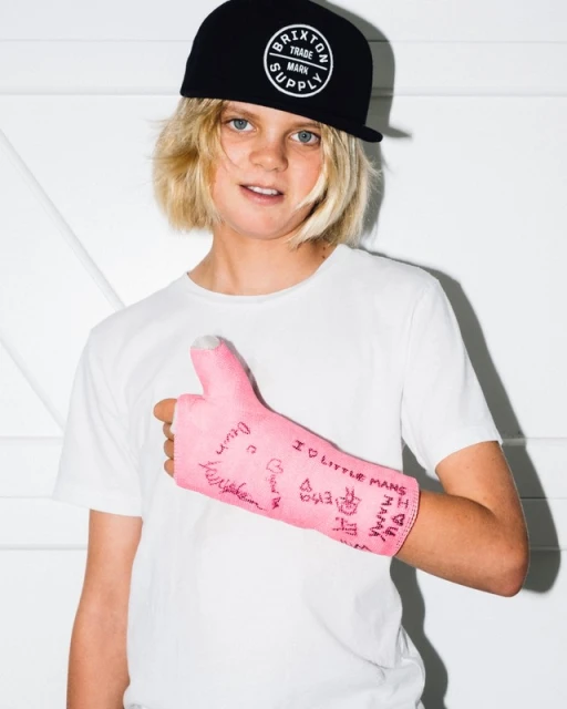 Young boy with blond hair, wearing a black cap and white t-shirt, poses with a pink arm cast covered in signatures and drawings.