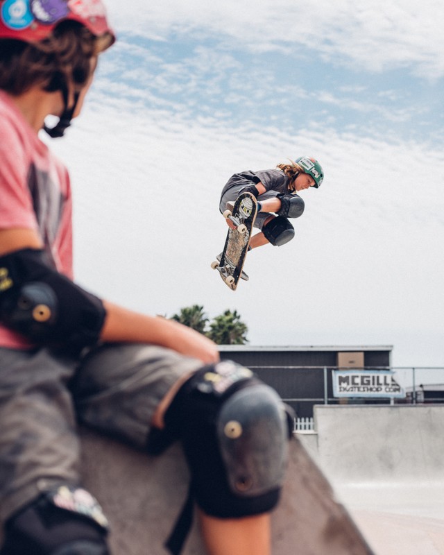 A skateboarder in mid-air performing a trick on a skateboard ramp, captured at a low angle with another person in the foreground.