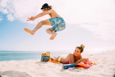 Man jumping in the air above a woman lying on a sandy beach under a clear sky.
