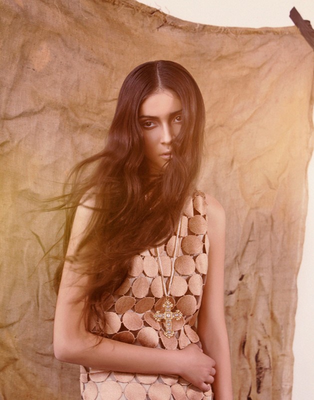 A young woman with long brown hair poses in a sleeveless dress with circular patterned details, wearing a cross necklace against a mottled brown background.