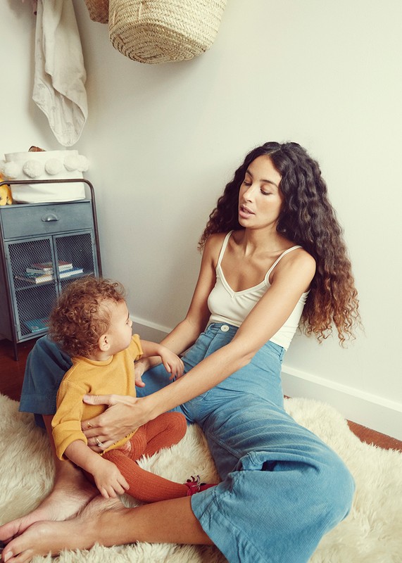 A mother and child are sitting on a furry rug in a brightly lit indoor space, with a metal cabinet and woven basket visible.