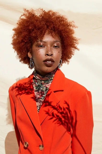 Portrait of a stylish Black woman with red curly hair, wearing an orange blazer and silver earrings, posing in front of a beige background.