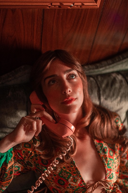 Woman lies down while holding a pink rotary phone with an upward gaze. She's wearing a patterned top in an indoor setting.
