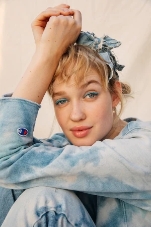 Close-up portrait of a smiling young woman with blonde hair, blue eyeliner, and a tie-dye sweatshirt against a neutral background.