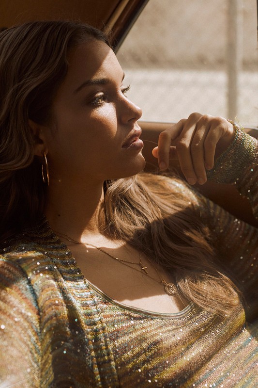 Profile of a woman with gold hoop earrings, wearing a gold, sparkling dress, sitting in a car and looking out the window with sunlight on her face.