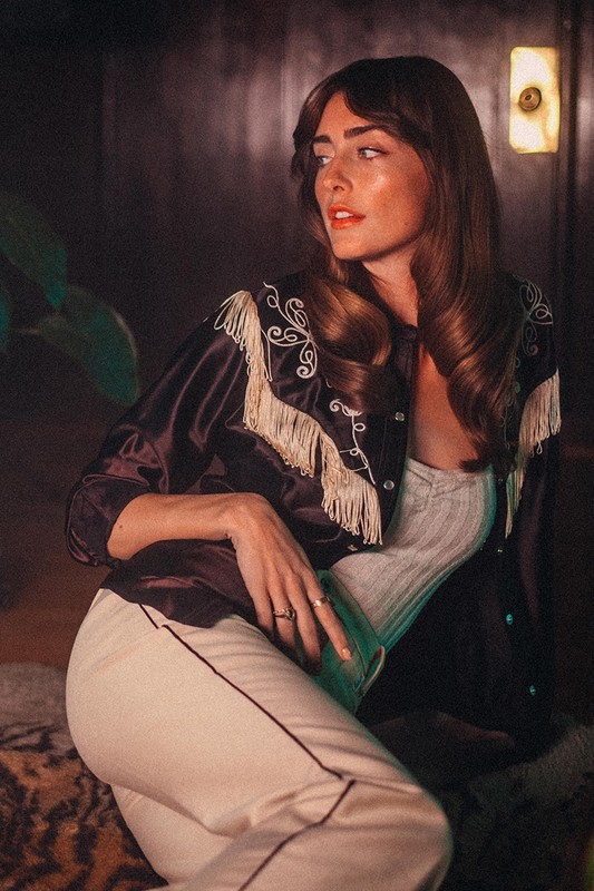 Attractive brunette woman with long wavy hair wearing a black fringe jacket and white pants posing indoors with a contemplative expression.