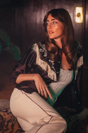 Attractive brunette woman with long wavy hair wearing a black fringe jacket and white pants posing indoors with a contemplative expression.