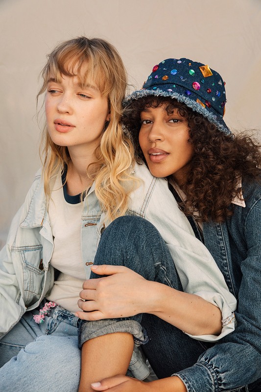 Two young women in denim jackets pose in a casual setting, one wearing a jeweled bucket hat, showcasing a blend of fashion and friendship.