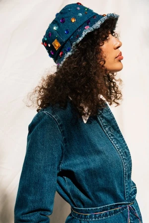 Woman in a denim dress and a jeweled denim bucket hat poses against a white background, shot in studio with diffused lighting.
