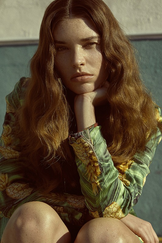 A young woman with wavy brunette hair poses indoors wearing a green patterned top, resting her head on her hand in a portrait.