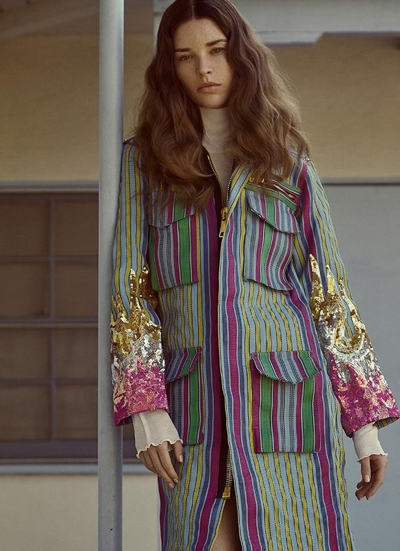 A young adult woman with long brown hair poses in a striped, embellished coat with a neutral background, fashionable and stylish.