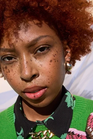 Close-up portrait of a Black woman with freckles, short red curly hair, and a green patterned sweater, shot by hair stylist Erika Parsons.
