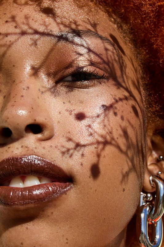 Close-up portrait of a woman with auburn hair, brown glossy lips, and freckles. Her face is partially covered by shadows of foliage.