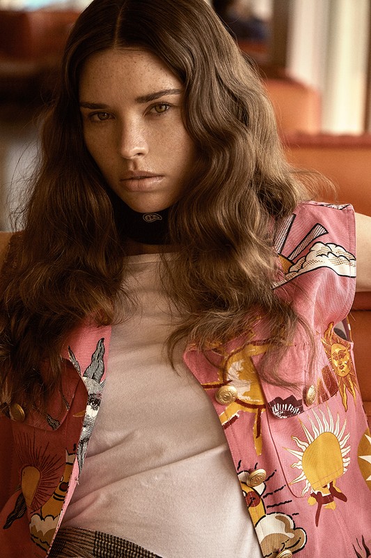 A young woman gazes forward wearing a white top and pink patterned vest, set against a warm, soft-focus interior background.