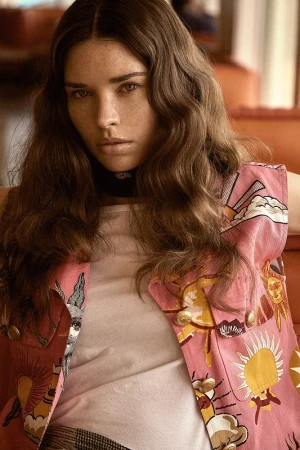 A young woman gazes forward wearing a white top and pink patterned vest, set against a warm, soft-focus interior background.