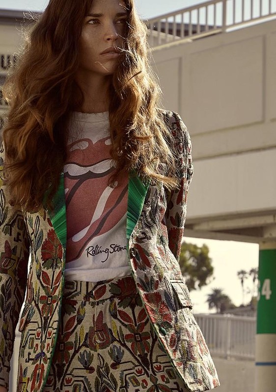 A young woman with long, wavy brown hair poses in an outdoor setting. She wears a printed blazer and matching skirt. The Rolling Stones logo adorns her t-shirt.