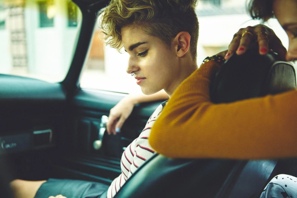 A young woman with blonde, wavy hair sits in the driver's seat of a car, with a passenger behind her, captured in a medium shot.