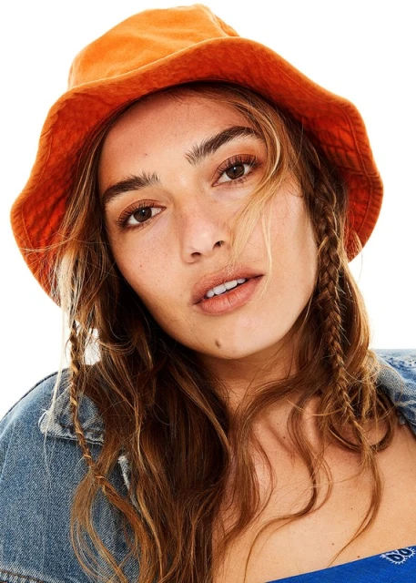 Close-up studio shot of an adult woman with fair skin wearing an orange bucket hat and a denim jacket, against a white backdrop.