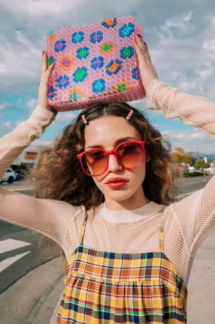 Young woman with curly hair wearing sunglasses and a plaid dress holds a beaded purse above her head against a blue sky backdrop.