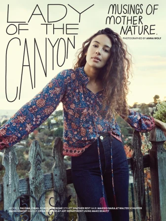 Woman with long curly hair wearing a floral print blouse leaning against a wooden fence overlooking hills