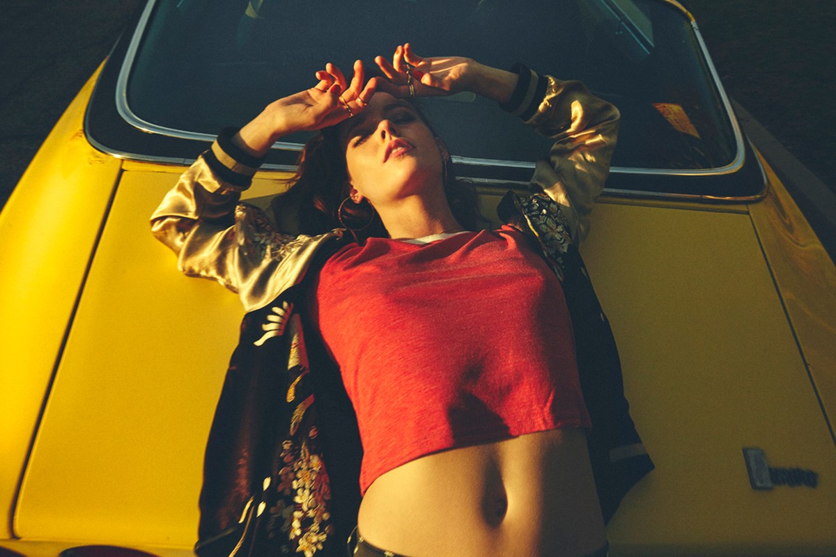 A woman in a red shirt and patterned bomber jacket lies on the hood of a yellow car, with her hands raised above her head.