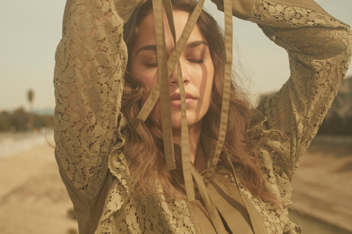 Adult female with her eyes closed wearing a green lace jacket, with tan ribbons in front of her face, outdoors in natural light.
