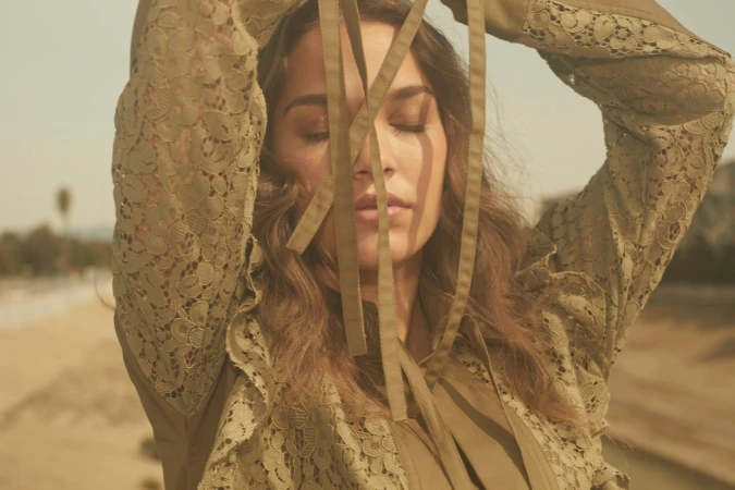 Adult female with her eyes closed wearing a green lace jacket, with tan ribbons in front of her face, outdoors in natural light.