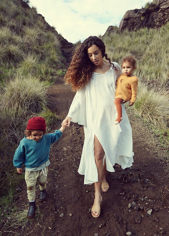 Woman with long curly hair wearing a white dress walking outdoors on a dirt path, holding a young child's hand and carrying another.