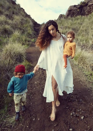 Woman with long curly hair wearing a white dress walking outdoors on a dirt path, holding a young child's hand and carrying another.