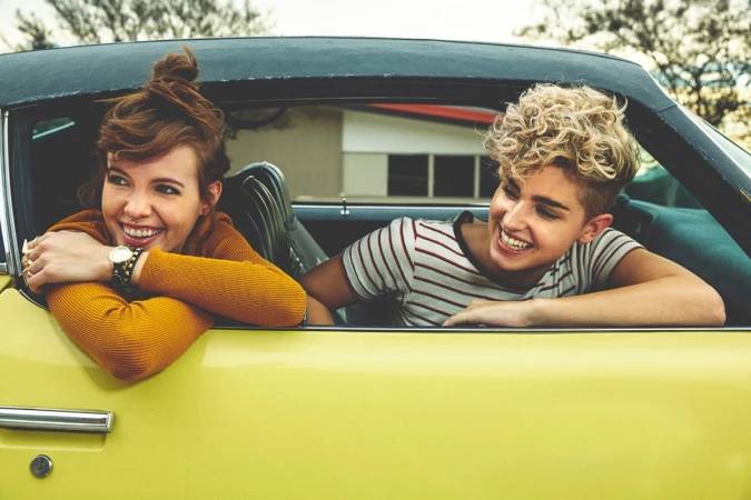 Two smiling young women enjoy a sunny day in a vintage yellow convertible. They exude happiness and carefree vibes during a road trip.