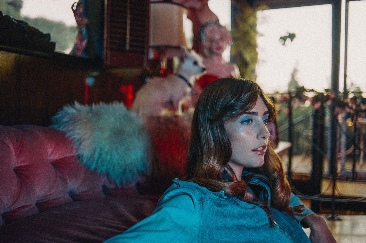 An adult female sits on a pink velvet couch inside an elegant interior, accompanied by a small white dog in the background.
