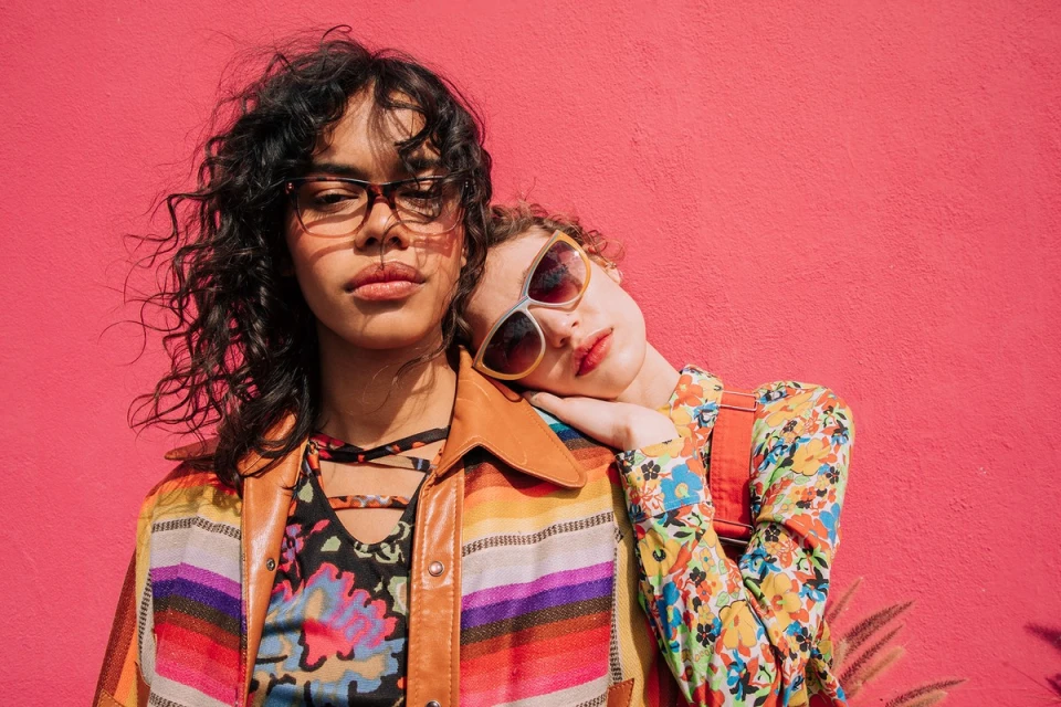 Two women pose against a bright pink wall. One woman rests her head on the other's shoulder, wearing colorful floral print clothing.