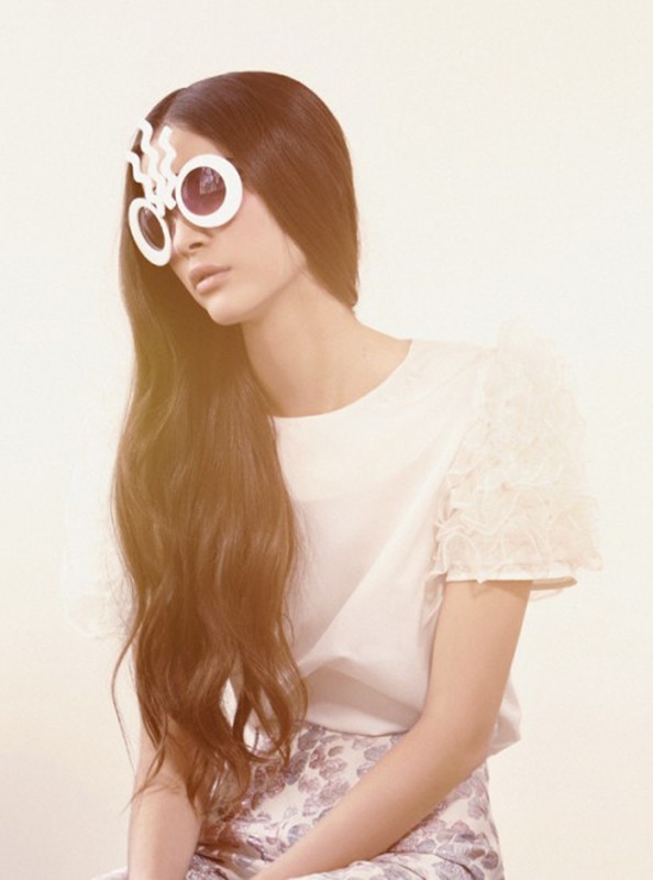 Fashionable woman poses with eccentric eyewear, wearing a white top and floral skirt against an off-white studio backdrop.
