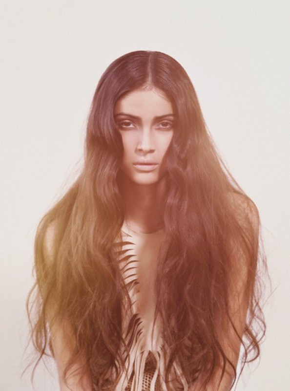 Portrait of a woman with long, dark hair and a serious expression against a white background, with a fringed top.
