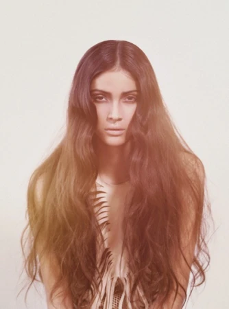Portrait of a woman with long, dark hair and a serious expression against a white background, with a fringed top.
