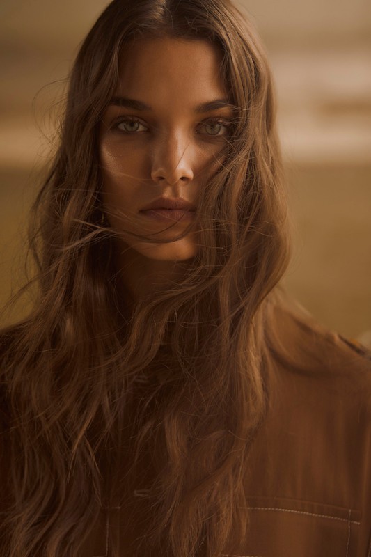 Close-up portrait of a woman with long brown hair, looking directly at the camera in a warm, neutral color palette, in an indoor setting.