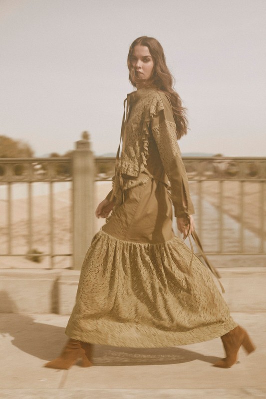 Fashionable adult woman in olive green dress and jacket walking on a stone walkway. Shot at eye-level, outdoors on a sunny day.
